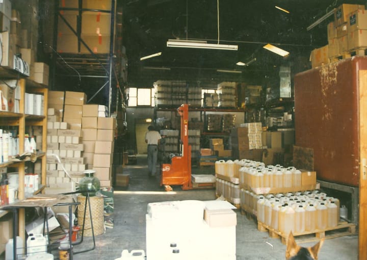 L’entrepôt des origines : rayonnages de cartons, palettes de bidons et transpalette manuel.